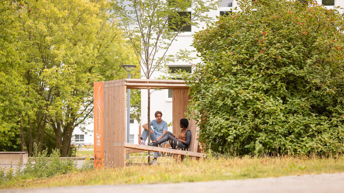 Foto von 2 Studierenden, die in einem Holzwürfel, der mit Sitzgelegenheiten ausgestattet ist, sitzen und sich unterhalten. In der Mitte des Würfels, ist ein Baumgepflanzt.