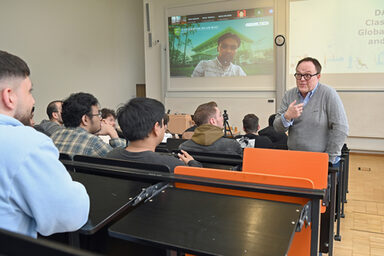 Eine männlich gelesene Person steht in einem Hörsaal und spricht engagiert zu einer Gruppe Studierenden. Auf der Leinwand ist digital eine weitere Person zugeschaltet.